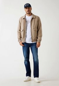 Giacca beige con zip e due tasche frontali, indossata sopra una t-shirt bianca, abbinata a jeans blu e sneakers bianche, completata da un cappellino navy.
