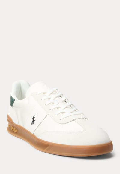 Biela nízka teniska s podrážkou z gumičky, čiernym logom Polo na boku, zelenou pätovou slučkou a bielymi šnúrkami, naklonená doľava na čistom pozadí.