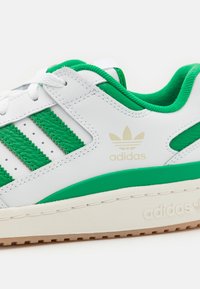 Sapatilha branca com detalhes verdes e três riscas verdes. Apresenta um cabedal de couro texturizado, perfurações para respirabilidade e uma sola de borracha.