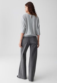 Femme aux cheveux bruns frisés portant un pull léger gris et un jean large gris foncé, se tenant de dos contre un fond uni.