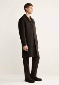Cappotto di lana marrone scuro con rever a punta, chiusura a un bottone, due ampie tasche anteriori a toppa, abbinato a pantaloni e scarpe scure coordinati.