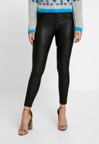 Pantalons en faux cuir noirs avec des détails côtelés et des coutures angulaires, associés à un pull gris clair et des talons en imprimé serpent.