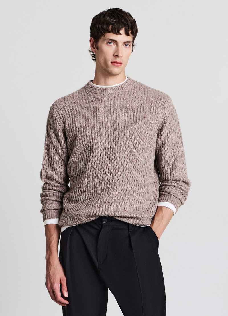 Jeune homme debout, une main dans la poche, portant un pull côtelé beige par-dessus une chemise blanche et un pantalon noir sur un fond uni.