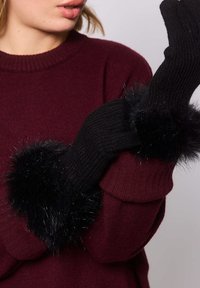 Mujer que lleva guantes de punto negros con puños esponjosos ajustando uno de los guantes, vestida con un suéter burdeos, frente a un fondo sencillo.