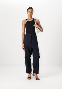 Haut sans manches noir avec un détail de bretelle, associé à un pantalon cargo taille haute bleu marine avec des accents de boutons et de poches. Tient un sac à main blanc.