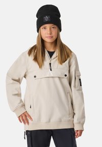 Giacca color crema con collo con zip, tasche laterali e dettaglio logo in evidenza. Indossata con berretto di maglia nero. Tessuto liscio.