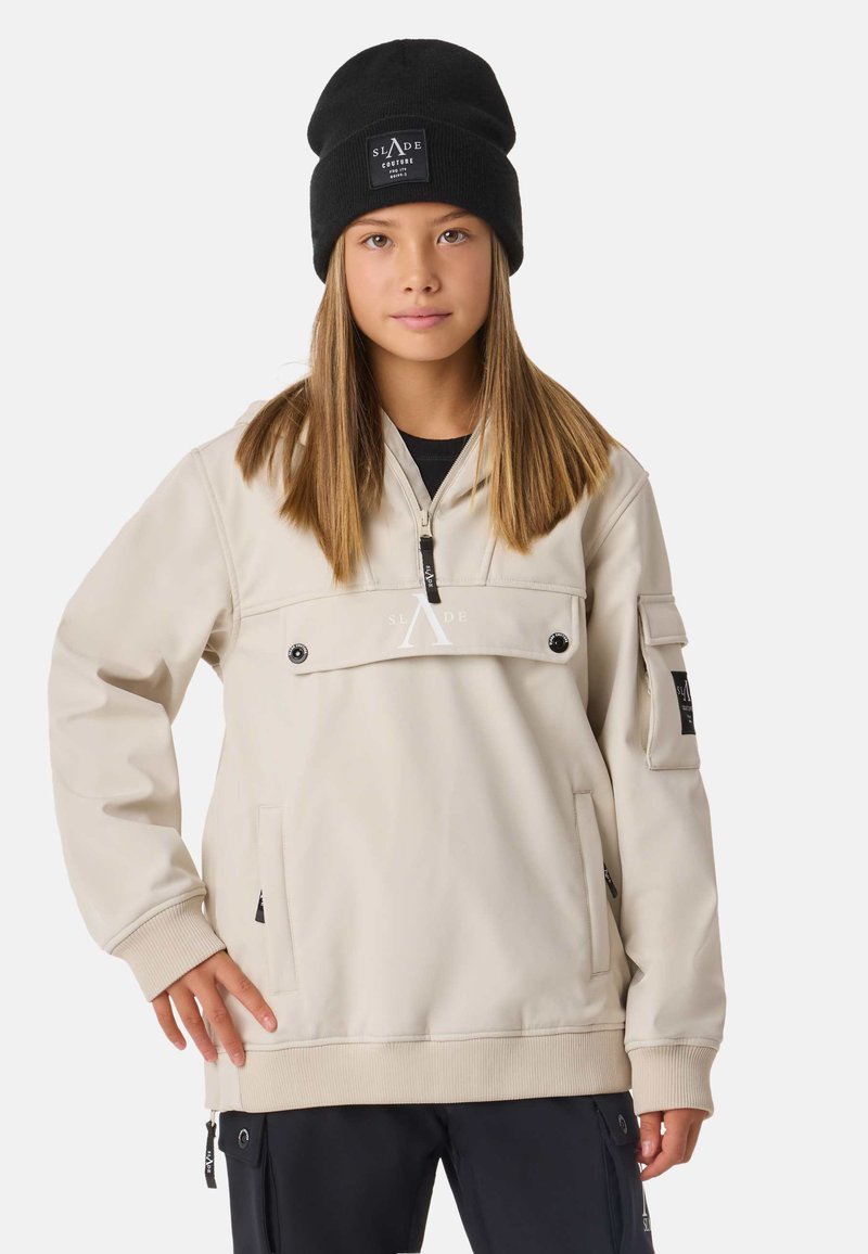 Giacca color crema con collo con zip, tasche laterali e dettaglio logo in evidenza. Indossata con berretto di maglia nero. Tessuto liscio.