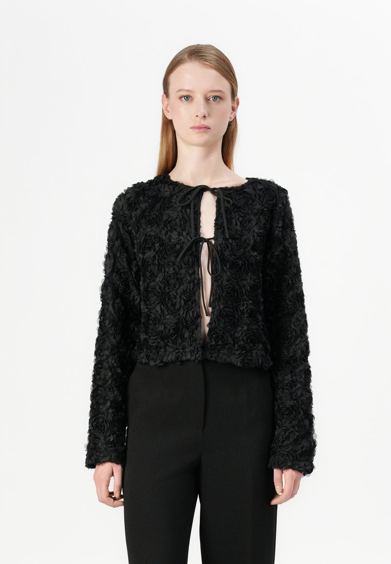 Veste noire en tissu floral texturé avec de longues manches et un col rond. Elle dispose d'une fermeture à lien sur le devant et d'un design court.