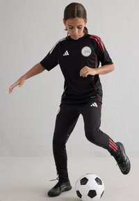 Melna sporta krekls ar sarkaniem akcentiem un balto logotipu, apvienots ar melniem biksēm, kuru malās ir trīs sarkanas līnijas. Melni futbola apavi un futbola bumba.