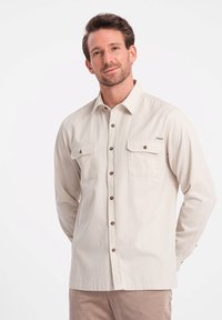 Lichtbeige langemouwhemd met knoopsluiting aan de voorkant, twee borstzakken, bruine knopen en een label aan de linkerkant. Soepele textuur.