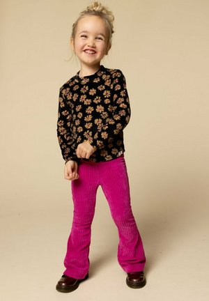 Lachend jong meisje met blond haar in een bun, gekleed in een zwarte bloemetjestop, felroze broek en bruine schoenen, staand tegen een beige achtergrond.