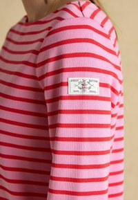 Zbliżenie na różowo-czerwoną koszulę z długim rękawem w paski, z białą prostokątną etykietą na górnej części rękawa.