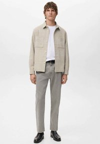 Cazadora de camisa para hombre de cuadros en beige claro, llevada sobre una camiseta blanca, combinada con pantalones gris ajustados y botines negros.