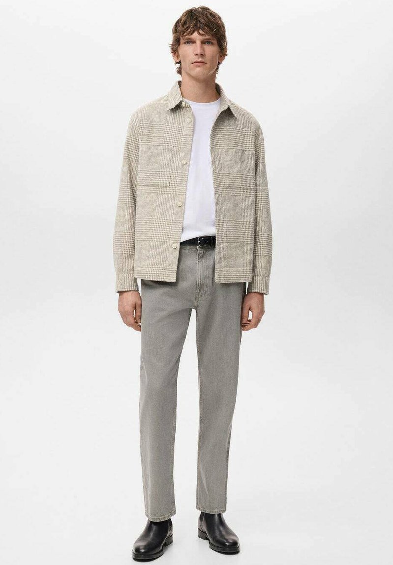 Herrenhemdjacke in hellem Beige mit Karomuster, getragen über einem weißen T-Shirt, kombiniert mit grauen Slim-Fit-Hosen und schwarzen knöchelhohen Stiefeln.