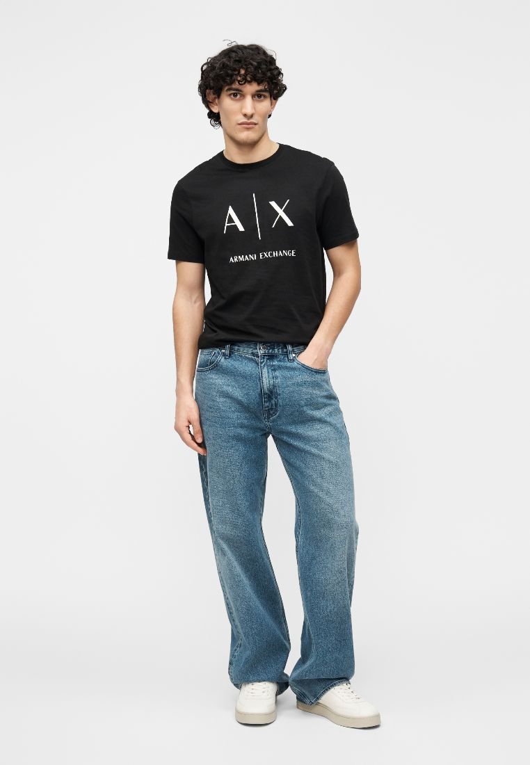 T-shirt en coton noir avec logo blanc "A|X ARMANI EXCHANGE". Jean denim bleu clair à coupe décontractée. Baskets blanches, posées sur un fond uni.
