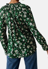 Kvindeligt iført en grøn blomstrede bluse med lange ærmer og plisseringer på den øverste ryg, kombineret med blå jeans.