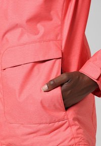 Korallfärgad jacka i en texturerad tygkvalitet, med en framficka och en hand som vilar i fickan. Ärmen har en ärmknapp.