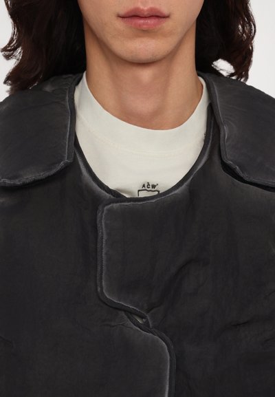 Veste noire texturée avec un grand col et une fermeture à boutons, superposée à une chemise à col rond de couleur claire avec un logo.