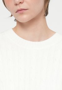 Prim-plan al feței inferioare și gâtului unei persoane care poartă un pulover alb din tricot cu model de cablu și un decolteu rotund cu dungi.