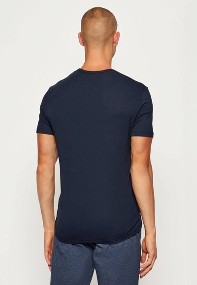 Άνδρας με ξυρισμένο κεφάλι στέκεται γυρισμένος με την πλάτη, φορώντας ένα απλό, σκούρο μπλε κοντομάνικο T-shirt και γκρι παντελόνι μπροστά σε λευκό φόντο.