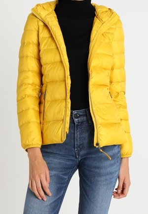 Kobieta w żółtej pikowanej kurtce na czarnej bluzce i niebieskich dżinsach, stojąca z rękami swobodnie opuszczonymi wzdłuż ciała.