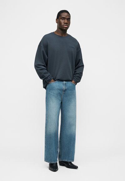 Jeune homme portant un sweat-shirt foncé surdimensionné, un jean bleu délavé à jambes larges, des bottines noires, debout les mains dans les poches sur fond blanc.
