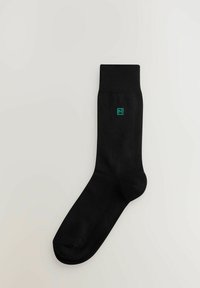 Sort bomuldssok med ribbet manchet og et lille grønt firkantet logo med bogstavet "N" nær anklen. Glat tekstur og standardform.