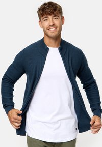 Cárdigan de punto azul marino con cremallera de longitud completa y cuello alto, llevado sobre una camiseta blanca, con puños acanalados y un acabado texturizado.