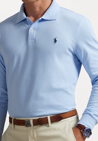 Mees, kes kannab helesinist pika varrukaga polo-särki, millel on broderitud logo, beežid püksid, pruuni nahkrihma ja sinist käekella, millel on nahkrihm.
