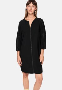 Schwarzes Kleid mit V-Ausschnitt, Frontreißverschluss und dreiviertel Puffärmeln. Auffällige weiße Steppnähte an den Nähten. Glatte Stofftextur.