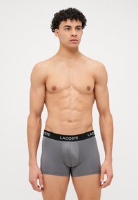 Sivi bokserice z črnim pasom Lacoste. Imajo tesen kroj in gladko teksturo. Poudarjeno šivanje ob šivih.