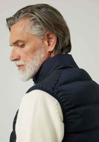 Chaleco de plumas azul marino con cuello alto, con textura acolchada. Lleva puesto un suéter de punto color crema, mostrando un diseño en capas.