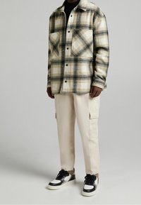 Hombre vistiendo una camisa de cuadros beige de botones, pantalones cargo crema y zapatillas en blanco y negro frente a un fondo gris liso.