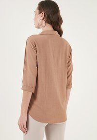 Brown, textured shirt with a collar and rolled sleeves, featuring a curved hem. No visible patterns or hardware details.
