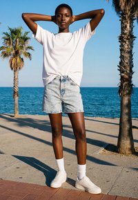 Young person wearing white t-shirt, denim shorts, and white sneakers stands with hands behind head near palm trees by the ocean under clear sky.