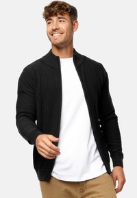 Cárdigan negro con cremallera y diseño de punto texturizado, con cuello alto y mangas ajustadas, llevado sobre una camiseta blanca.