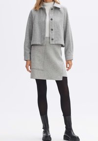 Femme portant une veste cropped grise à boutons et une jupe assortie avec des collants noirs et des bottines noires sur un fond uni.