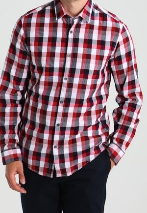 Mężczyzna ubrany w czerwoną, białą i czarną koszulę w kratę na guziki z długim rękawem oraz czarne spodnie, jedną ręką w kieszeni.