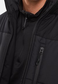 Sort isoleret jakke med høj krave, lynlåslommer og justerbar snoretræk hætte. Blød tekstur og synligt syet mønster.