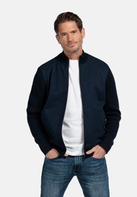Marine bomberjack met een ritssluiting aan de voorkant, gebreide manchetten en een gladde afwerking. Gecombineerd met een wit T-shirt en blauwe jeans.