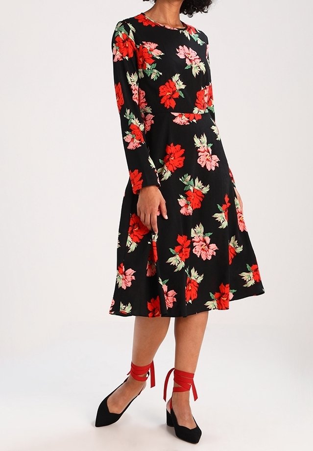 Vestido negro de flores con mangas largas, que presenta patrones de flores rojas y rosas, un corsé ajustado, falda acampanada y una textura de tela suave. Acentos de cinta roja en zapatos negros.