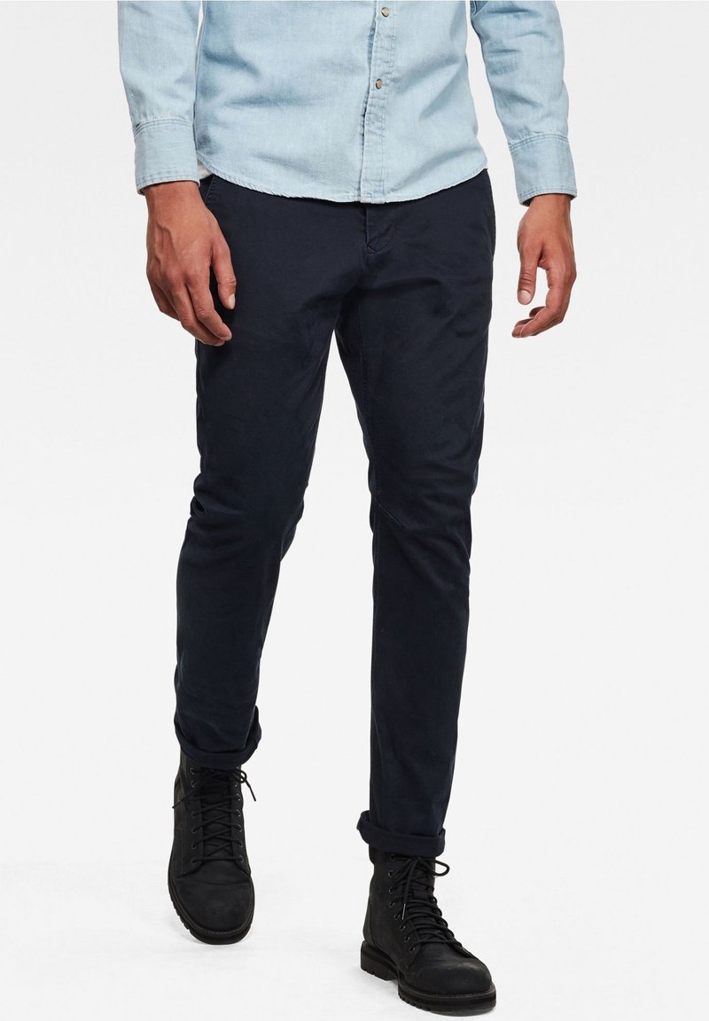 Homme portant un pantalon bleu marine foncé, une chemise à manches longues bleu clair avec des boutons et des bottes noires à lacets, debout devant un fond blanc.