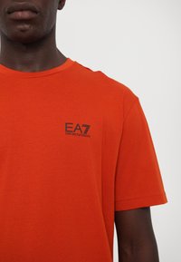 Tricou din bumbac portocalie strălucitoare, cu guler rotund, mâneci scurte și un logo proeminent EA7 Emporio Armani în negru pe piept.