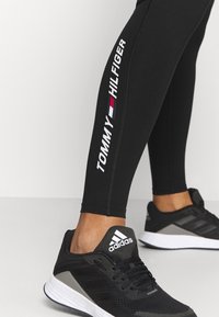 Czarne legginsy z białym logo "TOMMY HILFIGER" wzdłuż boku, połączone z czarno-szarymi trampkami Adidas z białą podeszwą.