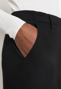 Sorte bukser med en glat tekstur og et tætsiddende design, med en sidelomme og en flad talje, parret med en hvid ribbet ærme.