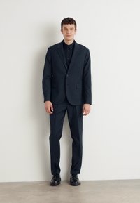 Abito scuro con giacca a tinta unita e pantaloni a gamba dritta, realizzato in tessuto liscio. Il modello indossa una camicia nera con bottoni e scarpe.