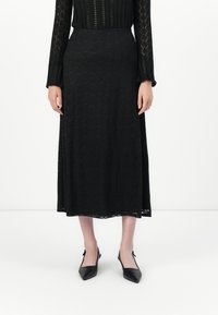 Kvinne iført en svart blonde langermet topp, matchende svart blonde midiskjørt, og svarte spissede slingback-flats, stående på hvitt gulv.