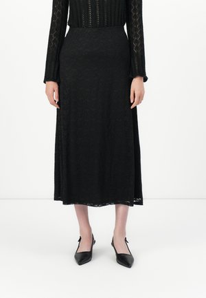 Kvinne iført en svart blonde langermet topp, matchende svart blonde midiskjørt, og svarte spissede slingback-flats, stående på hvitt gulv.