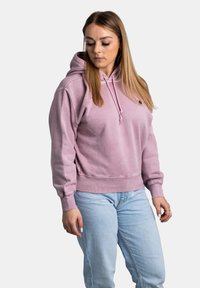 Sweat-shirt à capuche rose avec une poche kangourou, des poignets et un ourlet côtelés. Le tissu semble doux ; il est orné de cordons de serrage au niveau de la capuche et d'un détail logo.
