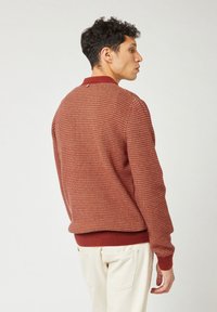 Mann, der seitlich steht und einen rostfarbenen gemusterten Pullover sowie off-white Hosen vor einem hellen einfarbigen Hintergrund trägt.